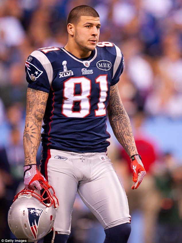 Football player Aaron Hernandez, in a blue jersey and silver pants, stands holding a silver helmet. The football player has a shaved head and arms covered in tattoos.